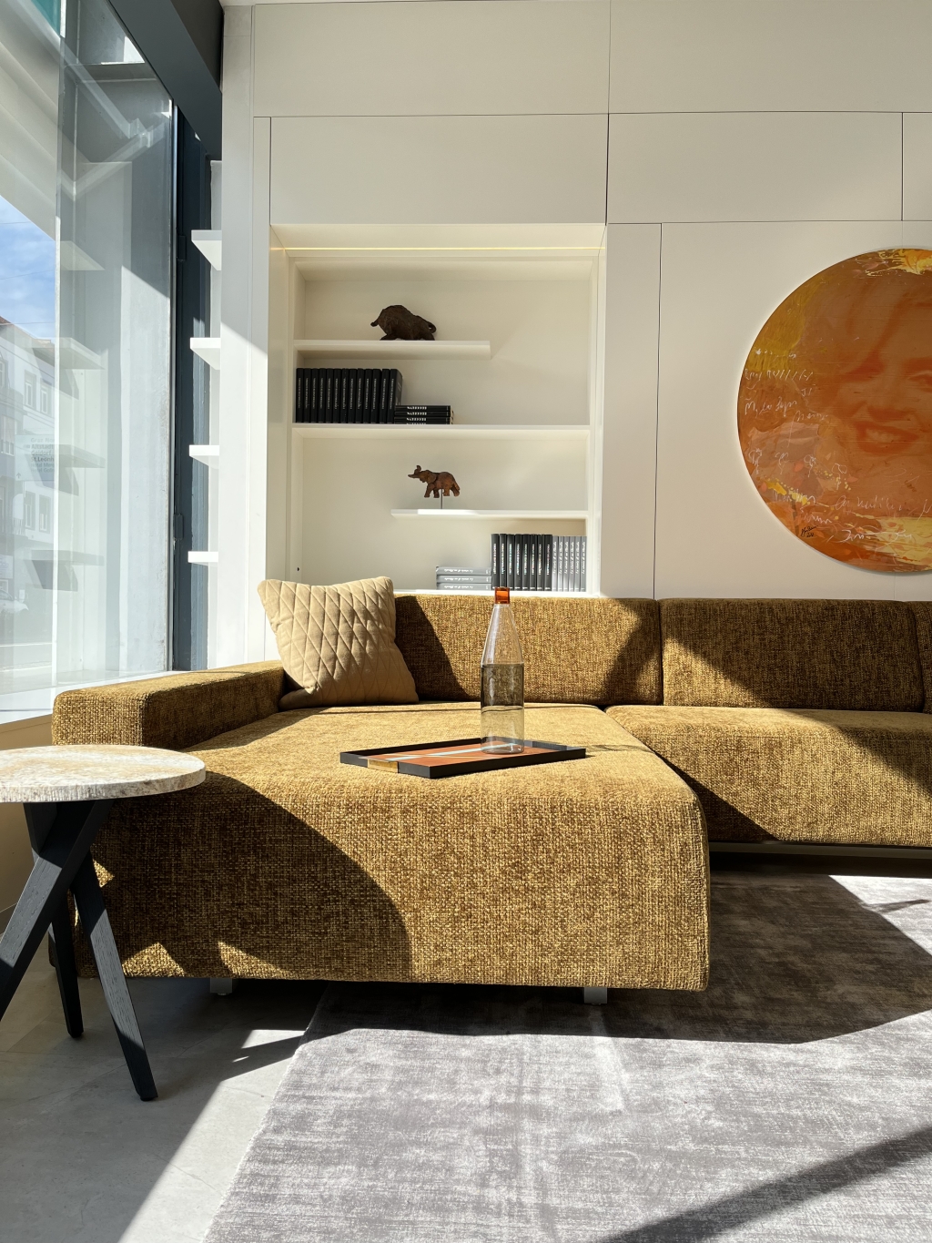 Ein gemütliches, modernes Wohnzimmer in natürlichem Sonnenlicht, mit einer stilvollen strukturierten Couch mit bequemen Kissen, einem eleganten Beistelltisch und schicken Regalen mit minimalistischem Dekor, ergänzt durch ein lebendiges kreisförmiges Kunstwerk an der Wand.