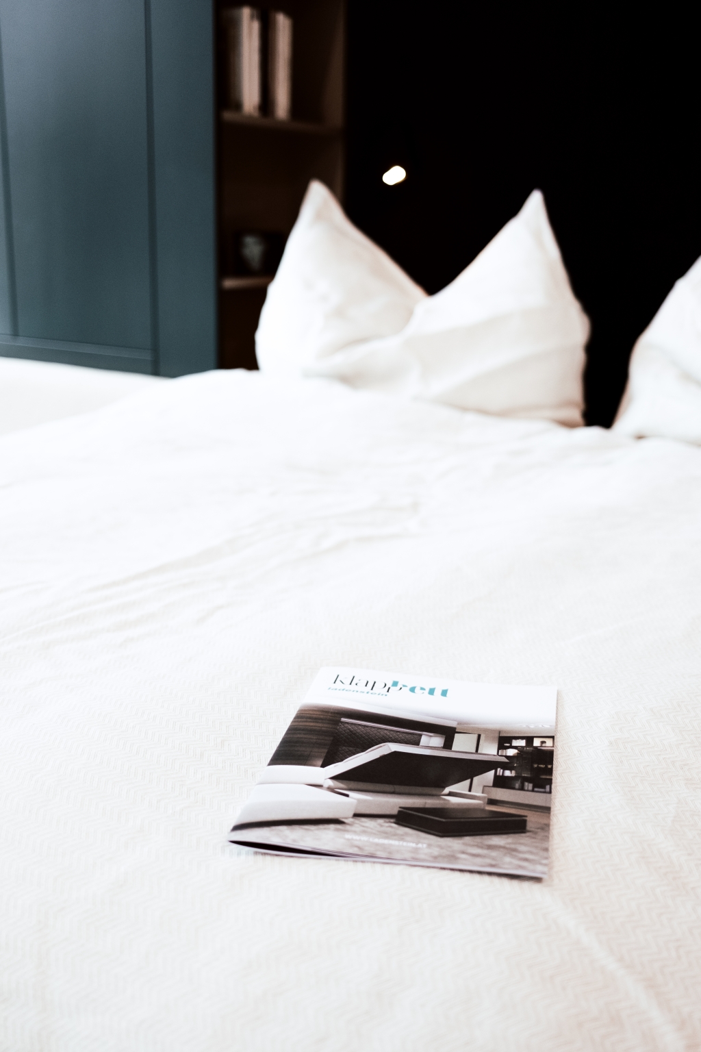Ein einladend gemachtes Bett mit frischer weißer Bettwäsche und flauschigen Kissen sowie eine in der Mitte ausgelegte Zeitschrift laden zu einer entspannten Lektüre in einer ruhigen Schlafzimmerumgebung ein.
