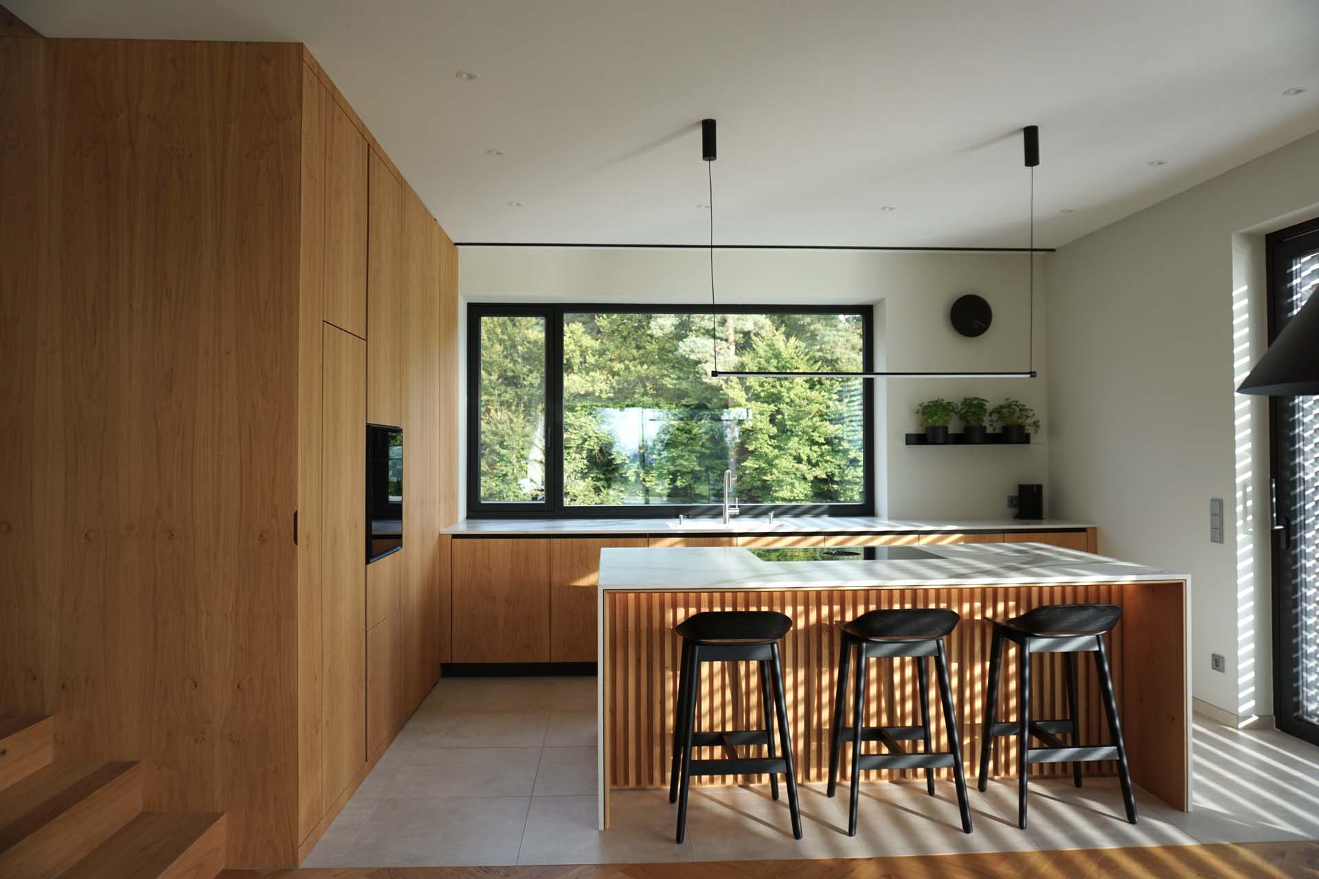 Eine moderne Küche mit eleganten Holzoberflächen, die durch große Fenster durch natürliches Licht beleuchtet wird, mit einer zentralen Insel mit Barhockern in einem Haus in der Steiermark.