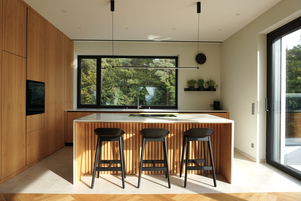Moderne Kücheneinrichtung mit eleganten Holzschränken, einer Kücheninsel mit Barhockern und einem großen Fenster mit Blick ins Grüne.