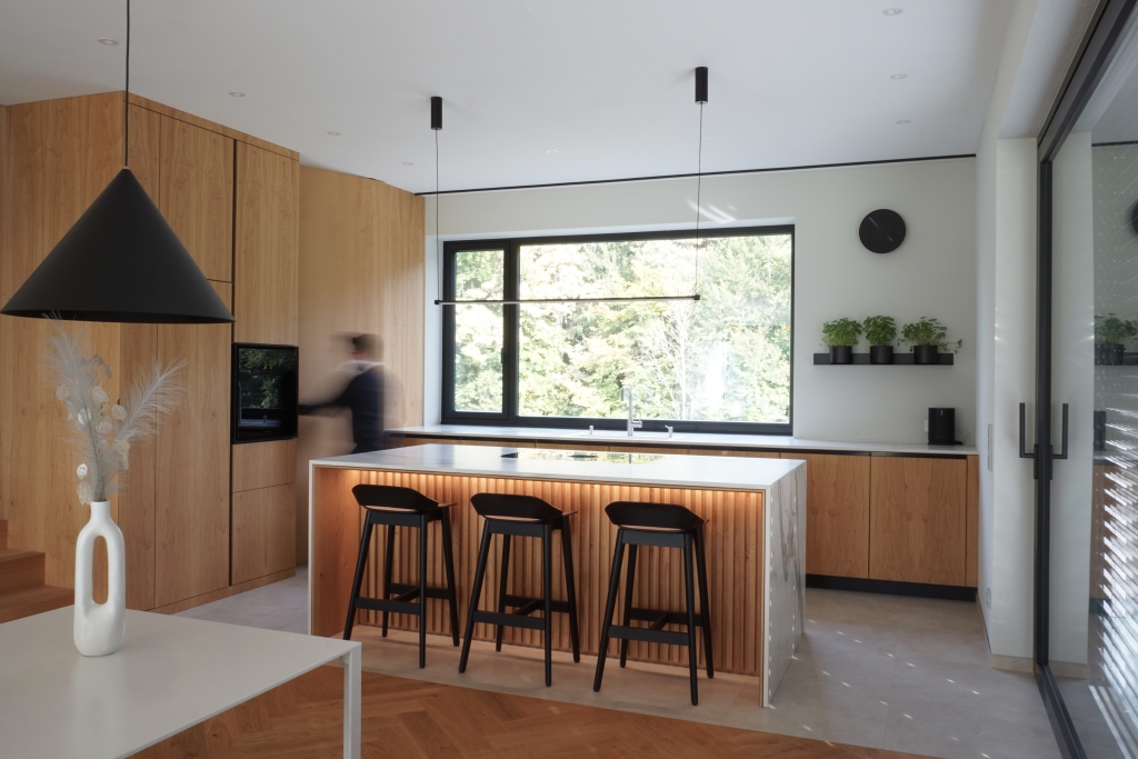 Eine elegante, moderne Küche mit Holzschränken, einer zentralen Insel mit Barhockern und großen Fenstern mit Blick ins Grüne.