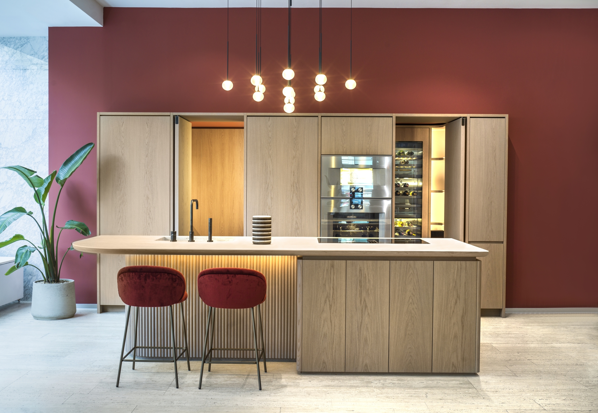 Modernes Kücheninterieur mit eleganter Holzoberfläche, einer Insel mit trendigen Barhockern und einer leuchtend burgunderroten Akzentwand, ergänzt durch stilvolle Hängelampen.