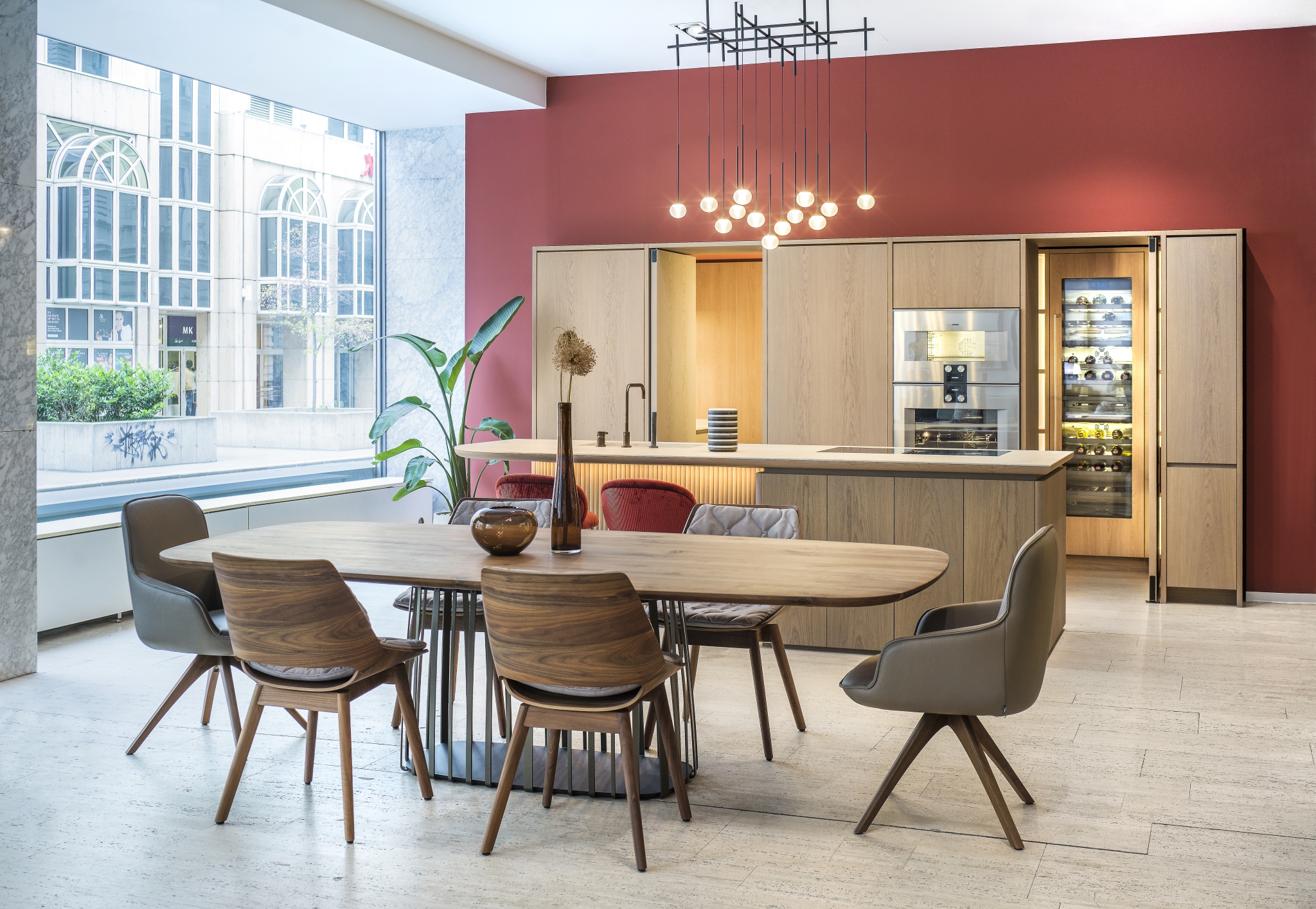 Eine moderne Küche und ein Essbereich mit einer kräftigen roten Akzentwand, Holzschränken, einem integrierten Weinkühlschrank und einem stilvollen ovalen Tisch, umgeben von gemischten Stuhldesigns unter einem eleganten Kronleuchter im Industriestil.