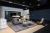 Moderne und schicke Wohnzimmerpräsentation mit einem weichen grauen Sofa mit Kissen, einem eleganten Couchtisch, einem stilvollen Sessel und eleganter Akzentbeleuchtung in einem eleganten Showroom-Ambiente.