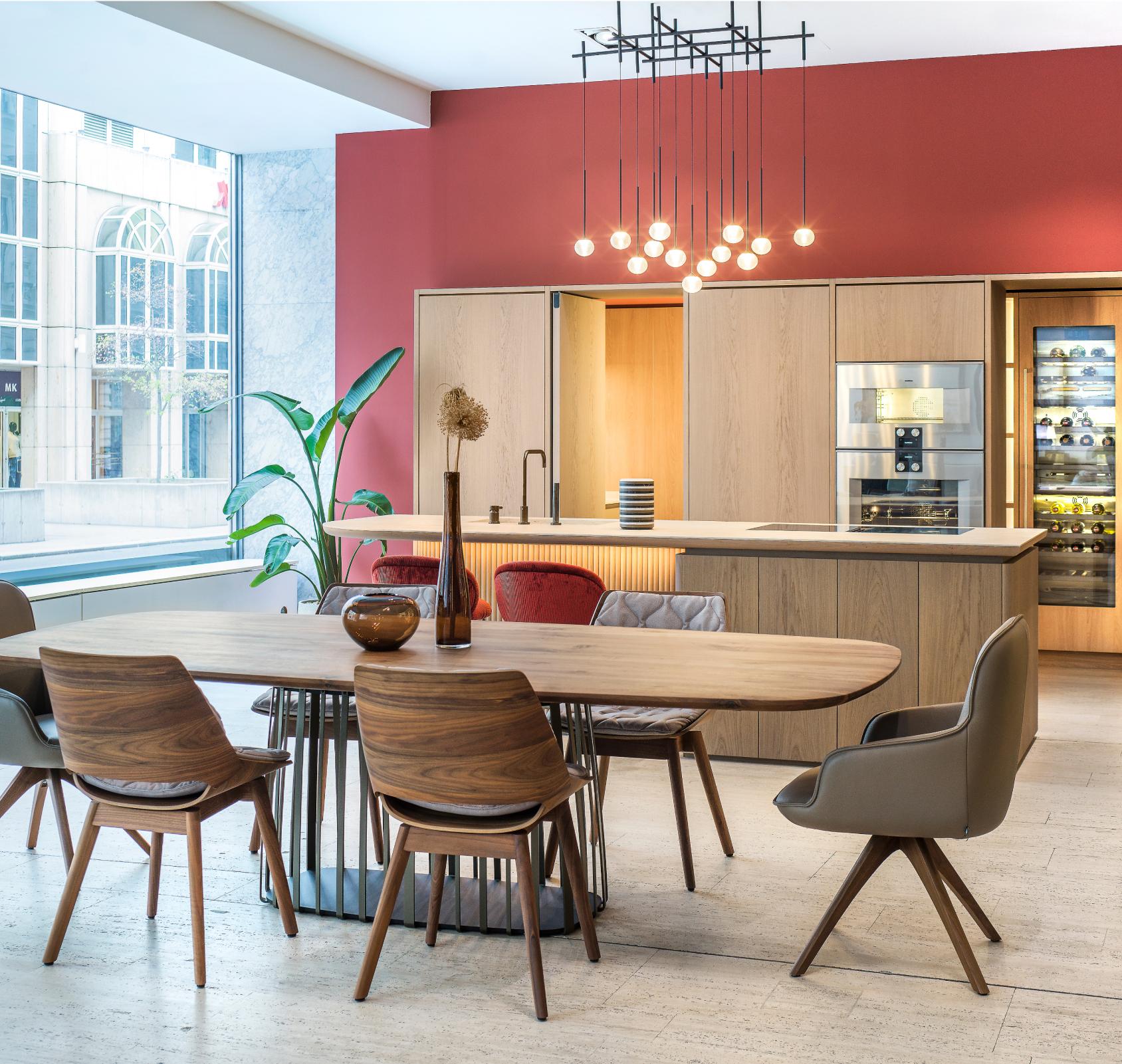 Ein moderner und stilvoller Küchen-Essbereich mit einem großen Holztisch, eleganten Stühlen und einer gut ausgestatteten Küchenzeile an einer warmen Terrakotta-Wand, ergänzt durch einen kunstvollen Kronleuchter und hohe Fenster, die den Raum mit natürlichem Licht durchfluten.