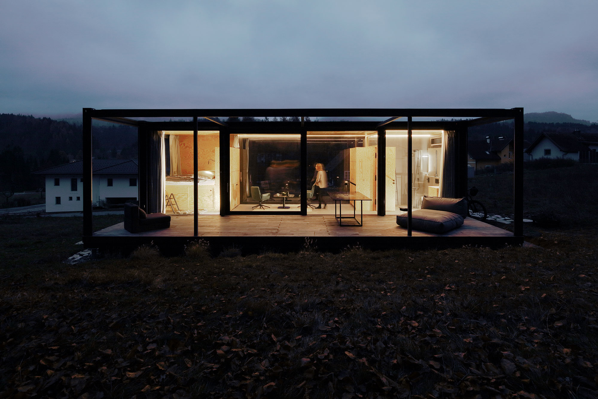Modernes, minimalistisches kleines Haus mit großen Glaswänden und einer eleganten schwarzen Rahmenstruktur, eingebettet in eine düstere Landschaft, mit einer sichtbaren Person im Inneren, die das gemütliche Interieur genießt.