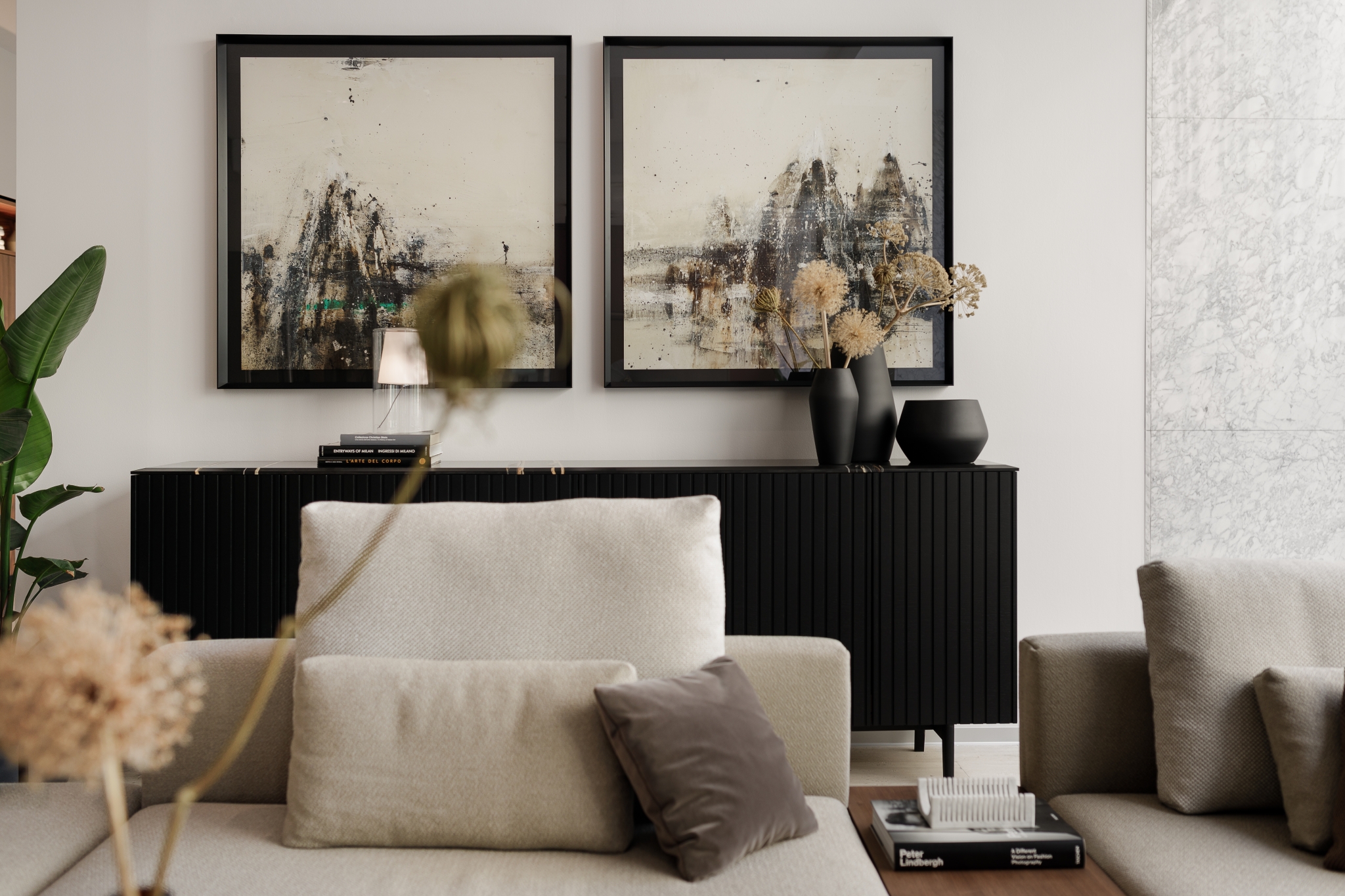 Moderne Wohnzimmereinrichtung mit neutraler Farbpalette und eleganten abstrakten Kunstwerken über einem eleganten schwarzen Sideboard, ergänzt durch gemütliche beige Sofas und geschmackvolle Dekorationselemente.